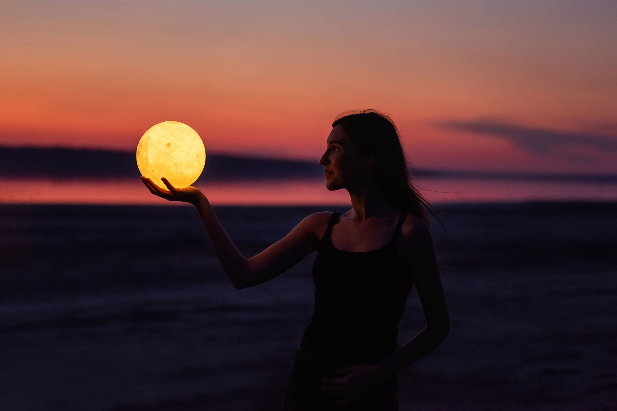 A young woman holds the full moon in her hands against the backdrop of a red sunset. Millennial blogger leads social media. Girl silhouette portrait. Astrology, Waxing Crescent. Copy space
